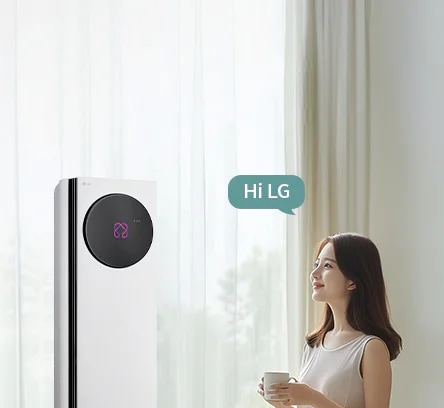 밝은 커튼이 드리워진 실내 공간에서 LG 휘센 AI 오브제컬렉션 뷰I 프로 에어컨이 왼쪽에 세워져 있고, 에어컨 본체 상단에 원형 블랙 디지털 디스플레이 화면에 현재 에어컨의 상태를 나타내는 아이콘이 보입니다. 오른쪽에는 컵을 들고 에어컨을 바라보는 여성이 창문을 통해 들어오는 자연광을 받으며 서 있습니다. 남성의 머리 위에는 Hi LG 라는 음성 인식 문구가 표시된 말풍선 형태의 그래픽이 보입니다. 이 이미지는 에어컨의 음성 인식 및 사용자 상호작용 기능을 실내 사용 환경과 함께 보여주고 있습니다.
