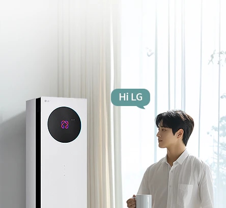밝은 커튼이 드리워진 실내 공간에서 LG 휘센 AI 오브제컬렉션 뷰I 프로 에어컨이 왼쪽에 세워져 있고, 에어컨 본체 상단에 원형 블랙 디지털 디스플레이 화면에 현재 에어컨의 상태를 나타내는 아이콘이 보입니다. 오른쪽에는 컵을 들고 에어컨을 바라보는 여성이 창문을 통해 들어오는 자연광을 받으며 서 있습니다. 여성의 머리 위에는 Hi LG 라는 음성 인식 문구가 표시된 말풍선 형태의 그래픽이 보입니다. 이 이미지는 에어컨의 음성 인식 및 사용자 상호작용 기능을 실내 사용 환경과 함께 보여주고 있습니다.
