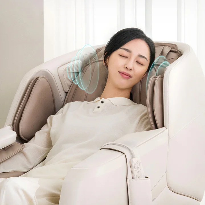 여성이 안마 의자에 앉아 눈을 감고 편안하게 쉬고 있습니다. 의자 양쪽에는 소리나 진동을 나타내는 듯한 파란색 원형 그래픽이 있습니다.